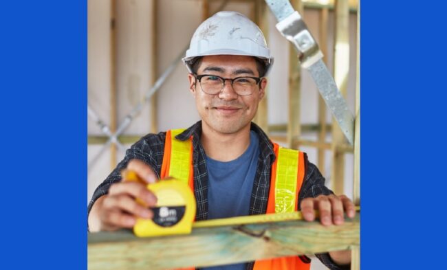 young man with safety helmet and glasses, measuring wood beam