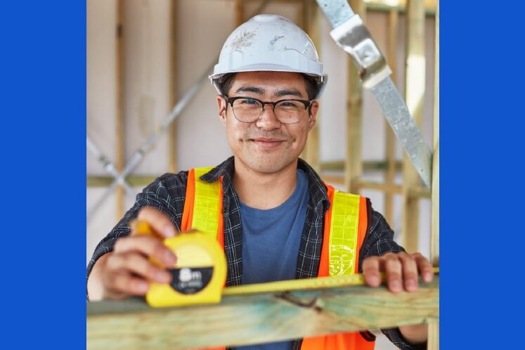 young man with safety helmet and glasses, measuring wood beam